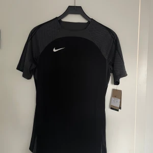 Svart fotbollströja från Nike - Snygg svart fotbollströja från Nike med korta ärmar och ett stilrent mönster på axlarna. Perfekt för träning eller match. Den har en vit Nike-logga på bröstet och är tillverkad i ett lätt och andningsbart material. Helt ny aldrig använd. 