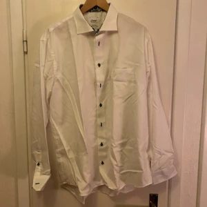 Vit skjorta från Oscar of Sweden - Säljer en stilren vit skjorta från Oscar of Sweden i regular fit. Skjortan har långa ärmar och svarta knappar som ger en snygg kontrast. Kragen har ett diskret mönster på insidan som ger en extra touch. Perfekt för en elegant look.