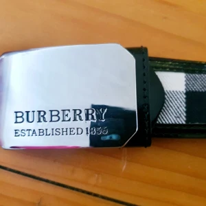 Burberry bälte - Burberry bälte/ skärp med klassiskt rutigt mönster i svart, beige och vitt. Bältet har ett stilrent spänne i silver med Burberry-logotypen ingraverad. Tyg/syntetskinn. Nästan nyskick, se bilder! 120 cm. Ej äkta.
