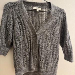 Kortärmad stickad kofta  - Säljer en grå stickad kofta från Moss med ett fint hålmönster.  Koftan är trekvarts/kortärmad men lite V neck💞