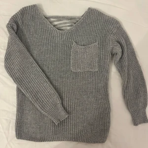 Grå stickad tröja med snörning - Säljer en mysig grå stickad tröja med en unik snörning i ryggen och en praktisk ficka framtill. 
