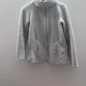 Tjock tröja fluffig  - Mysig grå teddyjacka med dragkedja och hög krage. Perfekt för kyliga dagar med sina långa ärmar och två praktiska fickor framtill. Jackan har en avslappnad passform och är superbekväm att bära.