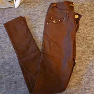 Bruna byxor med knappar - Snygga bruna byxor med en tight passform och dekorativa knappar. Perfekta för en stilren look. Byxorna har en klassisk design med fickor och är tillverkade i ett mjukt material.