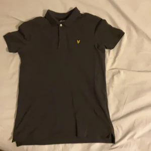 Svart pikétröja från Lyle & Scott - Säljer en stilren svart pikétröja från Lyle & Scott Junior. Tröjan har korta ärmar och en klassisk krage med knappar. Den är dekorerad med en gul logotyp på bröstet. Perfekt för en avslappnad och snygg look.