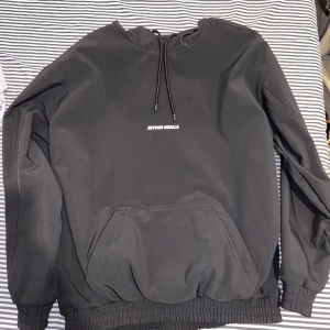 Svart hoodie från Beyond Medals. Beyond Medals vattenavvisande nightglow logo. - Säljer en svart hoodie från Beyond Medals med en stor ficka framtill och justerbar dragsko i halsen. Den har ett tryck på ryggen och ribbade muddar vid ärmslut och nederkant. Perfekt för en avslappnad stil.