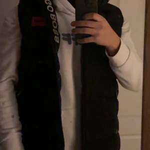 Svart dunväst från Hugo Boss - Snygg svart dunväst från Hugo Boss med logotyp på bröstet. Perfekt för lager-på-lager under kyligare dagar. Västen har en stilren design och passar bra över en hoodie eller tröja.