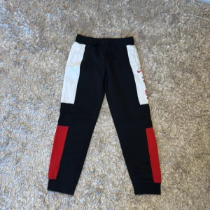 Svarta träningsbyxor från Nike - Snygga träningsbyxor från Nike i svart med vita och röda detaljer. Byxorna har en elastisk midja med snörning och är perfekta för sportiga aktiviteter. Nike-loggan syns tydligt på sidan.
