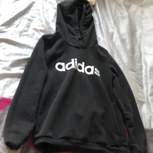 Svart hoodie från Adidas - Säljer en svart hoodie från Adidas med vit logga på framsidan. Tröjan har en klassisk design med huva och långa ärmar. Perfekt för en avslappnad stil.
