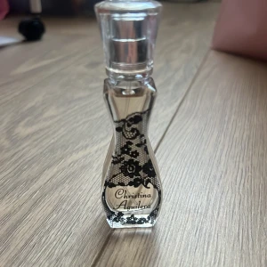 Parfym från Christina Aguilera - Elegant parfymflaska med en feminin design från Christina Aguilera. Flaskan är genomskinlig med ett svart spetsmönster och har en stilren, kurvig form. Perfekt för den som vill ha en doft med både stil och karaktär.