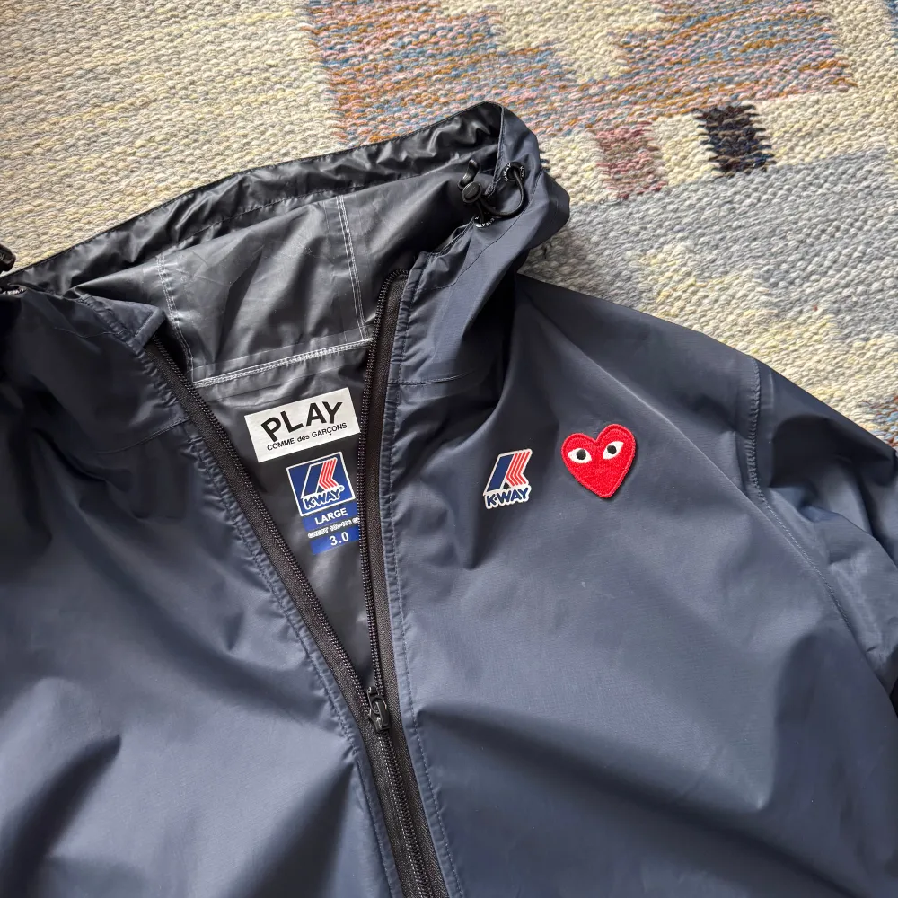 Säljer en stilren navy vindjacka från K-Way i samarbete med Comme des Garçons. Jackan har en dragkedja framtill och ett rött hjärta som detalj på bröstet. Perfekt för blåsiga dagar med sin lätta och funktionella design. Priset är inte hugget i sten 🪨. Takit.
