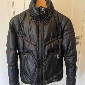 Dunjacka från J.Lindeberg - Snygg svart dunjacka med hög krage och dragkedja framtill. Jackan har två praktiska fickor med dragkedjor och quiltad design för extra värme. Perfekt för kyliga dagar och i skidbacken.