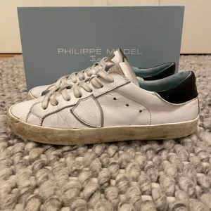 Vita sneakers från Philippe Model - Snygga vita sneakers från Philippe Model med svarta detaljer på hälen. Skorna har en klassisk design med snörning och är tillverkade i skinn. Perfekta för en stilren look, skorna har också en ”dirty look” Ny pris: 2800kr