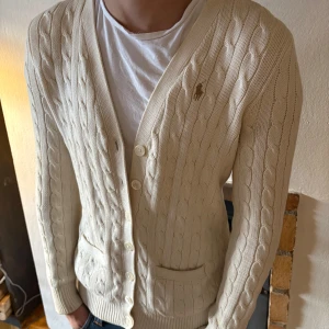 Beige stickad kofta från Ralph Lauren - Säljer en stilren beige stickad kofta från Ralph Lauren med kabelstickat mönster. Koftan har knappar framtill och en broderad logotyp på bröstet. Perfekt för en klassisk look.