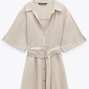 Beige klänning från Zara - Säljer en stilren beige klänning från Zara med knappar framtill och ett matchande bälte med gulddetalj i midjan. Klänningen har trekvartsärmar och en klassisk krage. Perfekt för en chic och avslappnad look.