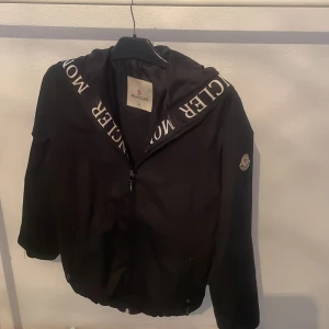 Svart jacka Moncler - Snygg svart jacka från Moncler med dragkedja och broderad logga på ärmen. Jackan har en huva med Moncler-text och långa ärmar. Perfekt för en stilren look.