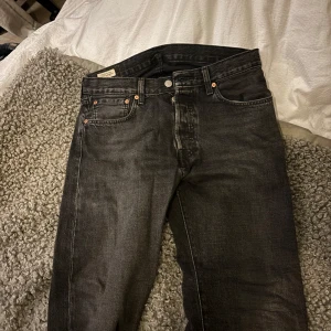 Svarta jeans från Levi's - Snygga svarta jeansbyxor från Levi's med klassisk femficksdesign och knappgylf. Perfekta för en stilren look. Märkeslappen i läder baktill ger en autentisk touch. Strl 32/32