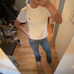 Vit t-shirt från Ralph Lauren - Snygg vit t-shirt från Ralph Lauren med det klassiska logotypbroderiet i blått på bröstet. Perfekt basplagg med rund hals och korta ärmar. Passar till alla tillfällen och är lätt att matcha med andra kläder.