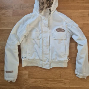 Vit vinterjacka  - Snygg vit vinterjacka med pälskantad huva och ribbade muddar. Jackan har dragkedja och knappar framtill samt praktiska fickor med lock. Perfekt för kalla dagar! ❄️