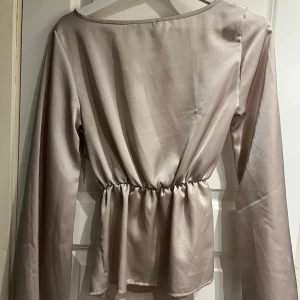 Beige långärmad blus från Boohoo - Elegant beige långärmad topp från Boohoo med en lätt glansig finish. Toppen har en elastisk midja som ger en smickrande passform och vida ärmar för en stilfull look.