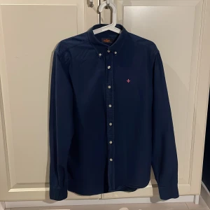 Mörkblå skjorta från Morris - Säljer en stilren mörkblå skjorta från Morris med broderad detalj på bröstet. Skjortan har långa ärmar och knappar framtill samt vid kragen. Perfekt för en elegant look.