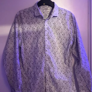 Vit blommig skjorta från Jack & Jones - Snygg vit skjorta från Jack & Jones med ett diskret blommigt mönster. Skjortan har långa ärmar och knappar framtill. Perfekt för en stilren look. Bara att fråga om frågor!