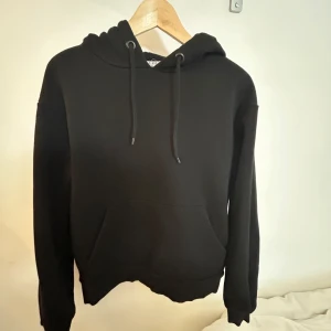 Svart hoodie med ficka - Säljer en klassisk svart hoodie med känguruficka och justerbar huva. Perfekt för en avslappnad stil och passar till det mesta. Skön och enkel att bära.