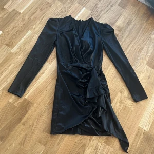 Svart omlottklänning - Elegant svart omlottklänning med långa ärmar och en v-ringad design. Klänningen har en stilfull drapering framtill och en asymmetrisk nederdel som ger en unik look. Perfekt för en kväll ute eller en speciell tillställning.