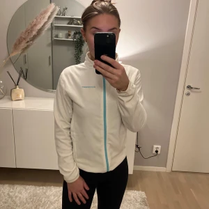 Vit fleecejacka från Everest - Säljer en vit fleecejacka från Everest med en snygg turkos dragkedja. Jackan har en hög krage och långa ärmar, perfekt för kyliga dagar. Den är mjuk och bekväm, idealisk för vardagsbruk eller utomhusaktiviteter.