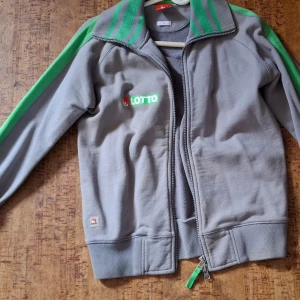 Grå och grön jacka från Svenska Spel - Säljer en grå jacka med gröna detaljer från Svenska Spel. Jackan har dragkedja framtill och långa ärmar med gröna ränder. Lotto-logga på bröstet och texten 'Vad drömmer du om?' på ryggen. Perfekt för en sportig look!