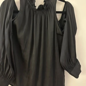 Svart blus med volangdetaljer - Elegant svart blus med volangdetaljer vid halsen och långa ärmar. Blusen har en lös passform och knappar framtill. Perfekt för en stilren look.