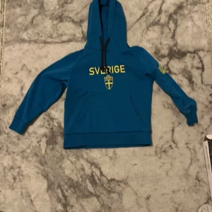 Blå hoodie från Admiral med Sverige-tryck - Säljer en blå hoodie från Admiral med texten 'Sverige' och en svensk flagga tryckt på framsidan. Tröjan har en praktisk huva och långa ärmar. Perfekt för att visa ditt stöd för Sverige!