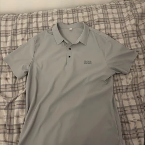 Grå pikétröja från Boss - Snygg grå pikétröja från Boss med klassisk krage och två knappar framtill. Perfekt för en stilren och avslappnad look. Boss-loggan är diskret placerad på bröstet. 