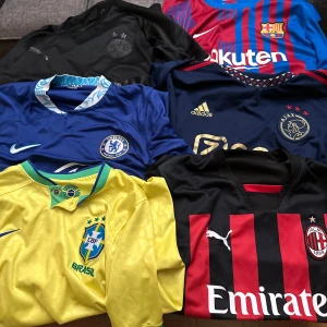 Fotbollströjor från olika klubbar och landslag - Samling av fotbollströjor från olika klubbar och landslag. Inkluderar en gul Brasilien-tröja, en blå Chelsea-tröja, en svart Borussia Dortmund-tröja, en röd och svart AC Milan-tröja, en blå och röd Barcelona-tröja och en mörkblå Ajax-tröja. Perfekt för samlare eller fotbollsfans!