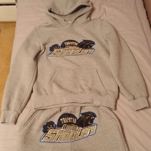 Grå hoodie från Trapstar - Säljer en grå hoodie från Trapstar med ett coolt 'London Shooters' tryck på framsidan. Den är perfekt för en avslappnad stil och har en stor ficka framtill. Huvan har dragsnören för justering. Superbekväm och perfekt för vardagsbruk!