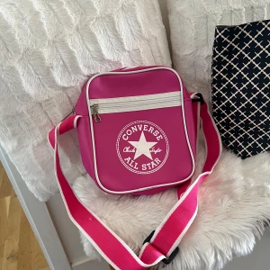 Rosa axelväska från Converse - Säljer en supercool rosa axelväska från Converse med det klassiska All Star-märket på framsidan. Väskan har en justerbar axelrem i rosa och vitt, och en praktisk dragkedja upptill. Perfekt för vardagsbruk eller när du vill lägga till en färgklick till din outfit!
