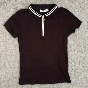 Svart kortärmad topp från FB Sister - Säljer en svart kortärmad topp från FB Sister i storlek XS. Den har en snygg dragkedja framtill och en krage med vita ränder. Perfekt för en casual look!