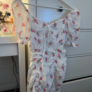 Blommig klänning  - Säljer en superfin blommig klänning från Shein i storlek S. Den är vit med rosa blommor och har en söt knytning framtill som går att justera. Klänningen har korta puffärmar och en dragkedja i ryggen. Perfekt sommarklänning🩷