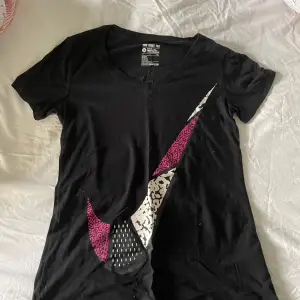 Svart t-shirt med en stor vit Nike-logga med rosa och svarta detaljer. T-shirten har en V-ringning.