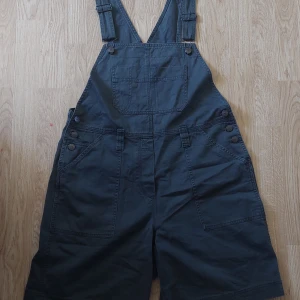 Snickarshorts - Hängselshorts/Mörkgråa snickarshorts köppta secondhand på Beyond retro i stockholm, storlek M/L banden kan justeras och knappar på sidan kan knäppas upp.