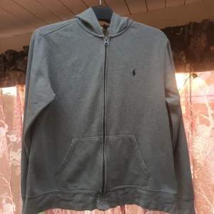 Grå hoodie från Polo Ralph Lauren - Säljer en stilren grå hoodie från Polo Ralph Lauren. Den har en dragkedja framtill och är tillverkad i ett mjukt bomullsmaterial. Perfekt för både höst och vår, med långa ärmar och en klassisk passform. Den ikoniska loggan är broderad på bröstet. Superbekväm och lätt att matcha med allt!