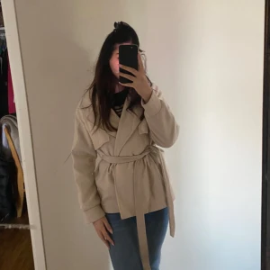 Beige kappa med bälte - Säljer en snygg beige kappa i storlek M. Den har ett bälte i midjan för en fin passform och långa ärmar. Perfekt för höst och vår, och den är i nyskick. Superchic och lätt att matcha med olika outfits!