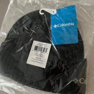 Svart mössa från Columbia - Säljer en helt ny svart mössa från Columbia. Den är fortfarande i förpackningen och har en enkel och stilren design. Perfekt för kalla dagar när du vill hålla dig varm och bekväm. Passar till det mesta i garderoben! ( Helt ny ) 