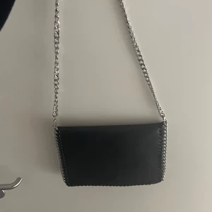 Svart axelväska med kedja - Säljer en snygg svart axelväska med en cool kedja som axelrem. Väskan är stilren och passar perfekt för både vardag och fest. Den har en enkel design utan extra detaljer, vilket gör den lätt att matcha med olika outfits.