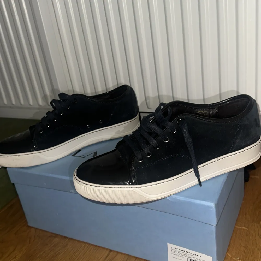 Säljer ett par stilrena blå  sneakers från Lanvin. De har en snygg glansig finish på tån och en vit sula som ger en cool kontrast. Skorna har klassisk snörning och är perfekta för både vardag . De är i bra skick 9/10. Är blå men ser ut som svarta . Skor.