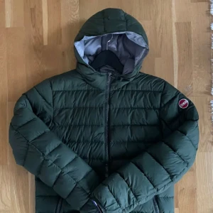 Grön pufferjacka från Colmar - Säljer en snygg grön pufferjacka från Colmar.  Varmt foder. Jackan har en dragkedja framtill och en huva för extra skydd mot väder och vind. Den kommer passa perfekt till vintern allra dagar. Den är i väl skicka men med två små hål som lätt ska gå och laga! Priset är sänkt från 1400 pga av skadan. Passar för dig mellan 165-175 cm