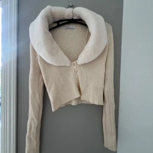Beige ribbad kofta med fuskpäls - Säljer en superfin beige ribbad kofta från Bershka. Den har en stor, fluffig fuskpäls-krage som verkligen gör den unik. Koftan är croppad och har knappar framtill. Perfekt för höst och vinter😇🤩 använd 2 gånger så är nästan i nyskick! 