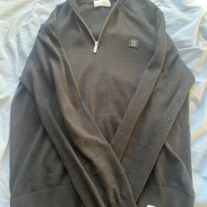 Svart halfzip från Les Deux - Säljer en stilren svart tröja från Les Deux med dragkedja och ribbad krage. Tröjan har en diskret logga på bröstet och är perfekt för en avslappnad look. Passar bra till både jeans och chinos.