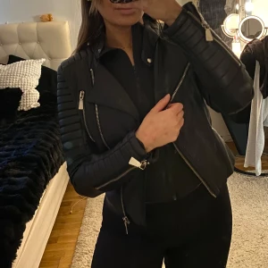 Svart skinnjacka med dragkedjor - Snygg svart skinnjacka med coola dragkedjedetaljer och bälte i midjan. Jackan har en klassisk bikerstil med quiltade axlar och långa ärmar. Perfekt för en edgy look!