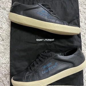 Svarta sneakers från Saint Laurent 42 - Snygga svarta sneakers från Saint Laurent med beige sula och blå logotyp på sidan. Skorna har klassisk snörning och är tillverkade i skinn. Perfekta för en stilren look. Är size 42 men passar 43 pris kan diskuteras 