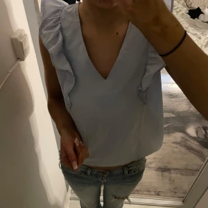 Ljusblå blus från Mango - Säljer en söt ljusblå blus från Mango med volangdetaljer vid axlarna. Blusen har en v-ringad design och är perfekt för en stilren look. Den är tillverkad i ett lätt och bekvämt material. Storleken är xl men passar mig bra som är en xs/s